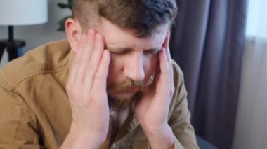 Evdeki koltukta oturup şakaklarına dokunarak baş ağrısından muzdarip 30 'lu yaşlardaki üzgün sakallı genç adama yakın durun. Migren, baş ağrısı, zihinsel stres, panik atak, gerginlik sorunu, akşamdan kalma konsepti