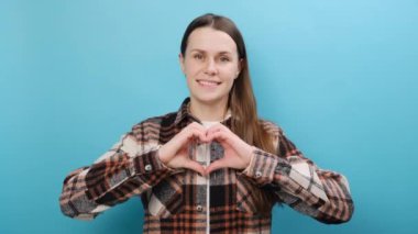 Çekici, pozitif genç bir kadının portresi, parmakları kurnazca gülümseyen bir kalp işareti gösteriyor. Cana yakın bayan, stüdyodaki mavi arkaplan duvarında izole edilmiş bir şekilde sevgi ve hayranlık gösteriyor.