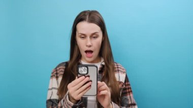 Üzüntülü genç kadın, günlük kıyafetler giyiyor cep telefonu küfrediyor. Kötü sahte haberler okuyor. Stüdyodaki pastel mavi arka plan duvarında izole edilmiş bir poz veriyor.