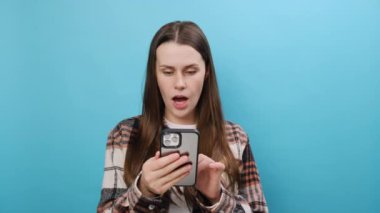 Heyecanlı, neşeli, beyaz bir kadın gündelik tişört giyiyor cep telefonunu işaret ederek cep telefonundan büyük kazanç haberi alıyor ve sade pastel mavi arka plan stüdyosunda izole bir şekilde poz veriyor.