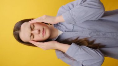 Hasta, yorgun, beyaz bir kadının dikey görüntüsü gündelik tişört giymiş ellerini baş masajı yapılan şakaklara koyuyor migrenden muzdarip, kötü hissediyor, sarı stüdyo arka plan duvarında izole edilmiş.