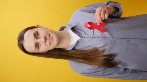 年轻的高加索女人拿着小红带的垂直镜头 看着相机 在工作室中将自己隔离在纯黄的背景墙之上 艾滋病毒认识概念 世界艾滋病日 — 图库视频影像