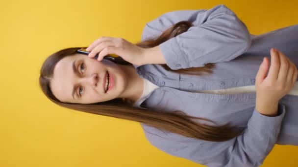 ハッピーな落ち着いた若い女性の垂直映像は 快適な会話をする携帯電話でシャツのトークを着て スタジオの黄色の背景の壁の上に隔離されています 人々のライフスタイルコンセプト — ストック動画