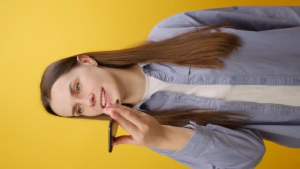 ソーシャルネットワークのオーディオメッセージで答える笑顔の若い女性の垂直ビデオは 黄色いスタジオの背景の壁に隔離されています スマートフォン仮想アシスタントをアクティブにするハッピーミレニアルレディ — ストック動画