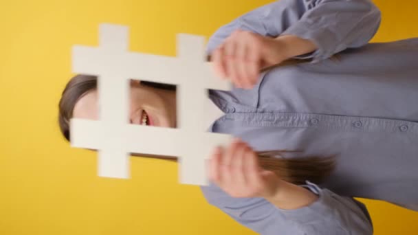 甜美的年轻女子面带微笑的垂直照片展示了Hashtag符号 正面看着相机 标注著名博客 病毒内容 穿着衬衫 与黄色工作室背景隔离 — 图库视频影像