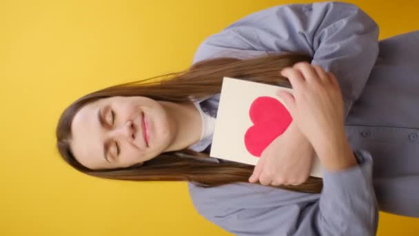 黄色の背景に隔離されたカメラを見ている幸せな描かれたカードを身に着けている美しい若い白人女性の笑顔の垂直映像 コンセプト愛とバレンタインカード — ストック動画