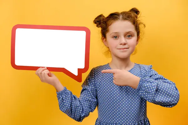 Portrait of pretty little girl pointing on speech bubble in ideas, opinion or vote, isolated on yellow studio background. Happy cute kid showing banner, paper or cardboard poster in speaker mockup