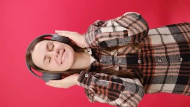 Gömlek giyen oldukça mutlu genç bir kadının dikey görüntüleri kulaklıkla mp3 müzik dinlerken, rahatlarken, stüdyodaki kırmızı arka plan duvarında izole bir şekilde poz verirken. Halk yaşam tarzı konsepti
