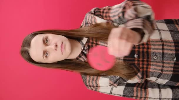 深刻な若いブルネットの女性25代の垂直映像は 不満の悲しい顔の絵文字を保持し スタジオの明白な赤い背景の壁の上に隔離されています コンセプト ストレス ネガティブな感情 — ストック動画