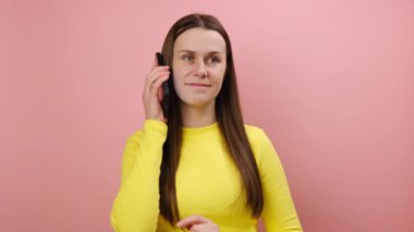 Neşeli, genç bir kadın cep telefonuyla arkadaşlarıyla konuşuyor, neşeli bir şekilde gülümsüyor, hoş sohbetler ediyor, sarı süveter giyiyor, stüdyodaki pembe arka plan duvarına izole edilmiş poz veriyor.