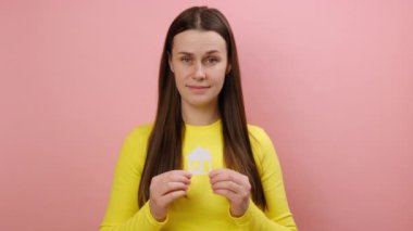 Yüzünde mutlu bir ifade olan beyaz bir ev tutan, sarı süveter giyen, stüdyodaki pembe arka plan duvarına izole edilmiş bir şekilde poz veren, beyaz bir kadın.