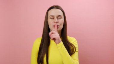 Gizli konsept. Güzel, kurnaz, gülümseyen genç bir kadının portresi. Gizli bir işaret, dudaklarına yakın bir parmak işareti, sarı süveter giyiyor. Stüdyodaki pembe renkli arka plan duvarına izole edilmiş poz veriyor.