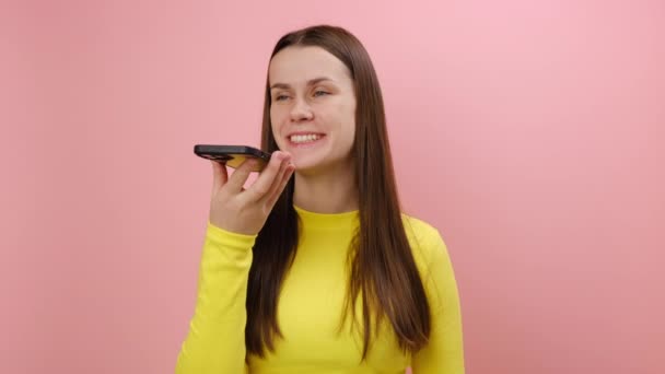 スマートフォンと話す幸せな若い女性 仮想アシスタントを使用して Aiインテリジェントテクノロジー 音声アプリケーション オーディオメッセージ セーターを着用し スタジオでピンク色の背景の壁に隔離されています — ストック動画