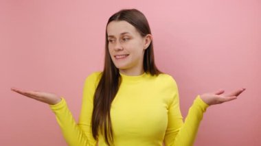 Kafası karışmış 25 'li yaşlarındaki genç bir kadının portresi, omuzlarını silkiyor, sarı kazak giyip stüdyoda pembe arka plan duvarına izole edilmiş bir şekilde poz veriyor.