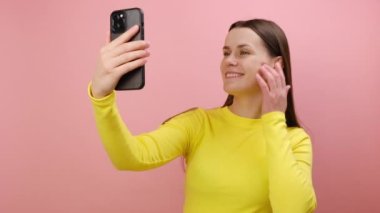Çekici, mutlu, genç bir kadın telefon kamerasıyla öpüşürken görüntülü konuşma ya da video bloğuyla iletişim kuruyor, sarı kazak giyiyor, stüdyodaki pembe renkli arka plan duvarına izole edilmiş bir şekilde poz veriyor.