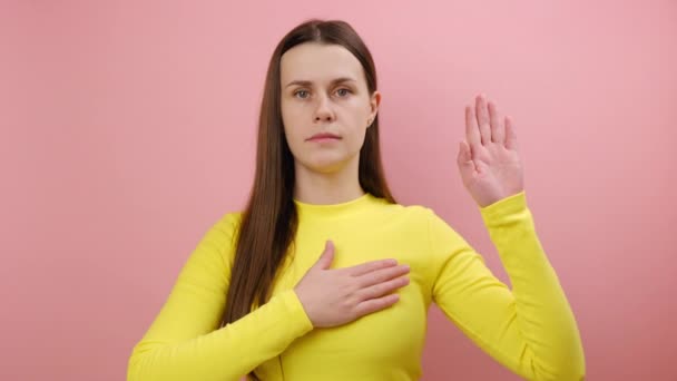 真剣な意識の若い女性 胸に手を保持し 別の誓いを立てます 信頼と正直 愛国心 黄色のセーターを着用 スタジオでピンク色の背景の壁の上に隔離されたポーズ — ストック動画