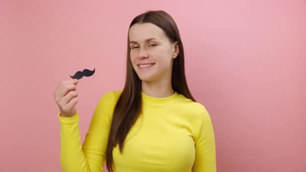 面白い遊び心のある若い白人女性は偽のペーパー口ひげを保ち カメラを見て幸せ お祝いの気分を持って 黄色いセーターを着てスタジオの明白なピンク色の背景の壁の上に隔離されたポーズ — ストック動画