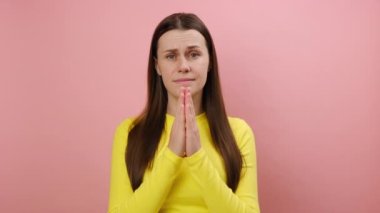 25 'lerinde genç bir kadının portresi dua ederken el ele tutuşuyor ve yalvaran yüz ifadesi, umut konsepti, sarı süveter giyme, pembe stüdyo arka planında izole edilmiş bir şekilde yardım istiyor. Lütfen, bana...