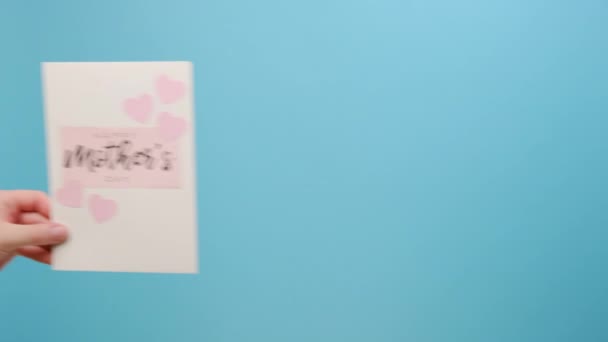 母の日のカードを小さなピンクのハートで保持している女性の近くに プロモーションコンテンツのコピースペースを持つスタジオの明白な青い背景壁に隔離されています 春休みのコンセプト — ストック動画