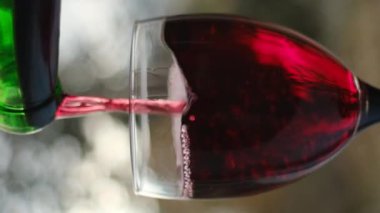 Güneşli bir günde, güzel bir arka planda, şişeden kırmızı şarap dökülüşünün dikey videosu. Şarap arıtma konsepti. Yavaş çekim