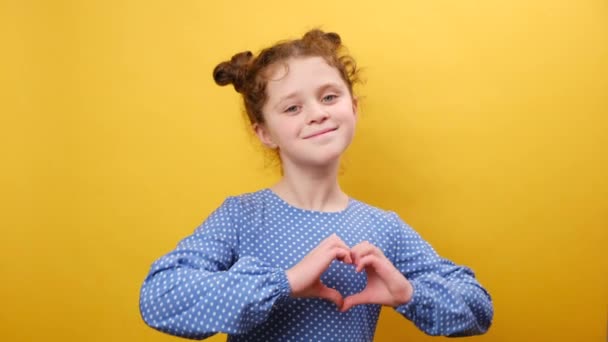 愛と慈善のコンセプト かなり幸せな小さな白人少女の肖像画 心のジェスチャーを示し カメラに微笑み 優しさを表現し スタジオで黄色の背景の壁の上に隔離された — ストック動画