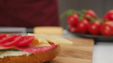Mutfaktaki ahşap tahtada taze kırmızı domatesli, salamlı, peynirli ve yeşil salatalıklı lezzetli sandviçler pişiren kimliği belirsiz genç bir kadın. Fast food konsepti