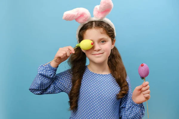 Portrait of playful cute happy little girl child in pink bunny ears holding near face easter eggs, smiling looking at camera, posing isolated over blue color background wall in studio. Holiday concept