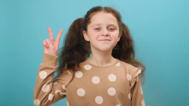 Vサインを示す魅力的なポジティブな少女の子供の肖像画 カメラを見て広く笑顔 広告のためのコピースペースを持つスタジオの明白な青い背景の壁の上に隔離されたポーズ — ストック動画