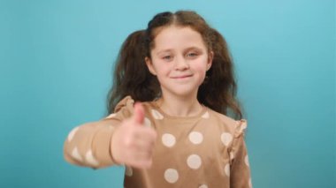 Şakacı, pozitif küçük kızın portresi başparmaklarını kaldırıyor ve kameraya samimi bir şekilde gülümsüyor. Stüdyoda mavi arkaplan üzerine izole edilmiş bir poz veriyor ve reklam için boş bir alan yaratıyor. Sevmek ve onaylamak