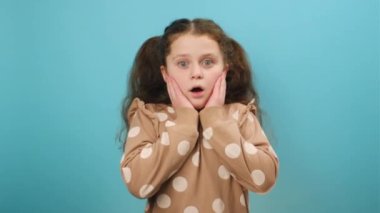 Omg. Ergen bir kızın portresi ağzını açıyor ve yüzüne dokunuyor, şok içinde kameraya bakıyor, stüdyodaki sade mavi arka plan duvarının üzerinde izole bir şekilde poz veriyor. İnsanlar duygusal yaşam tarzı konsepti