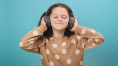 Büyük kablosuz kulaklıklarla müzik dinleyen, dans eden, kameraya mutlu bakan stüdyodaki sade mavi arka plan duvarında poz veren güzel küçük bir kızın portresi. Neşeli ruh hali