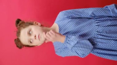 7-8 yaşlarındaki düşünceli küçük kızın elinde kırmızı soru işareti işareti, kafa karışıklığı konsepti olan dikey videosu stüdyodaki kırmızı arka plan duvarına izole edilmiş. Çocukluk yaşam tarzı konsepti