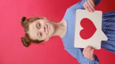 7-8 yaşlarındaki gülümseyen küçük kız çocuğunun dikey görüntüleri sosyal ağdaki kalp formuna benzer büyük bir işaret taşıyor, stüdyodaki kırmızı renk arka plan duvarında izole edilmiş şekilde duruyor. Çocukluk yaşam tarzı konsepti