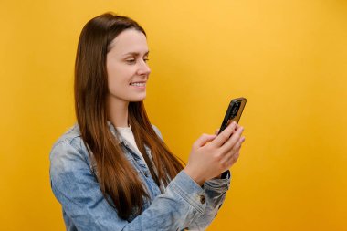 Gülümseyen genç kadın portresi akıllı telefondaki sosyal ağ iletileri, cep telefonundan memnun olan kullanıcı, kot ceket giyiyor, stüdyodaki sarı arka plan duvarında izole edilmiş poz veriyor.