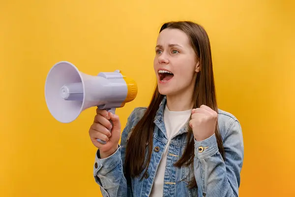 Portrait of angry young woman loudly screaming holding speaker in hand, strict boss yelling, raising fist, wearing denim jacket, isolated on yellow background wall in studio. Solving problem concept