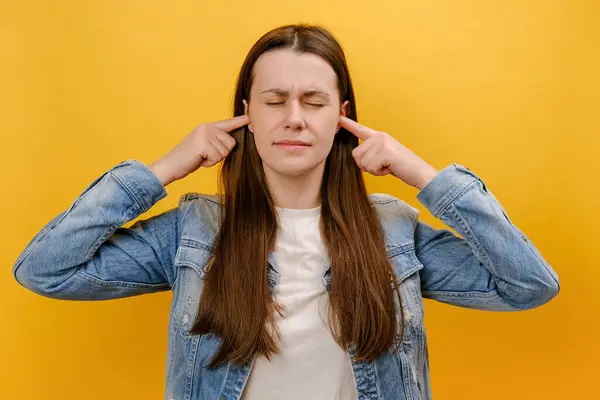 Portrait of nervous young woman standing and putting finger on ears and don\'t want to hear, nods her head, saying no, wearing denim jacket, posing isolated over yellow color background wall in studio