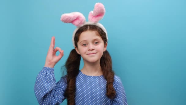 照片中漂亮的 面带微笑的十几岁以下小女孩身穿粉色兔子耳朵和休闲装 高兴地看着相机 用手指显示出不错的标志 在蓝色的工作室背景墙上摆出孤立的姿势 — 图库视频影像