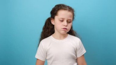 Üzgün küçük bir kızın portresi, depresif görünüyor, kasvetli suratlı bir çocuk, enerjisi yok, beyaz tişört giyiyor, stüdyoda mavi arka plan duvarının üzerine izole edilmiş pozlar veriyor.