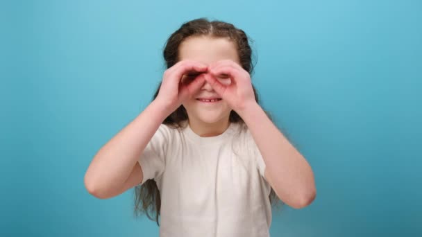 在演播室里 可爱的小女孩穿着白色休闲装 在蓝色的背景墙上摆出一副与世隔绝的样子 用双筒望远镜形状的手指看着外面 表达惊讶的形象 — 图库视频影像