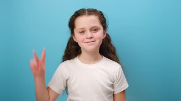 照片中可爱的小女孩双手空拳地展示自己的签名 为胜利做手势 为成功做手势 穿上白色休闲装 在工作室的蓝色背景墙上摆出孤身一人的样子 — 图库视频影像