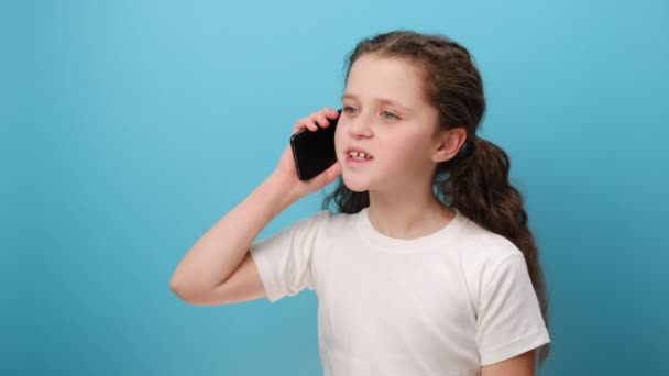 かわいい女の子の肖像画 携帯電話で話す 良い携帯電話 快適な子供のモバイルデバイスを使用するために快適 白のTシャツを着用 スタジオで青色の背景壁の上に隔離されたポーズ — ストック動画