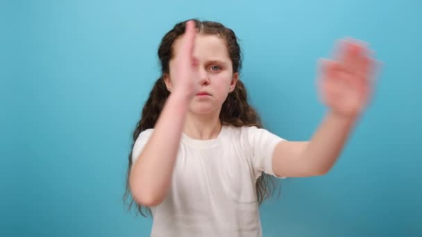 认真的小女孩交叉双手的画像 带着挑衅的目光看着相机 摆出停止姿势 禁止穿白色T恤 在工作室的蓝色背景墙上摆出孤立的姿势 — 图库视频影像