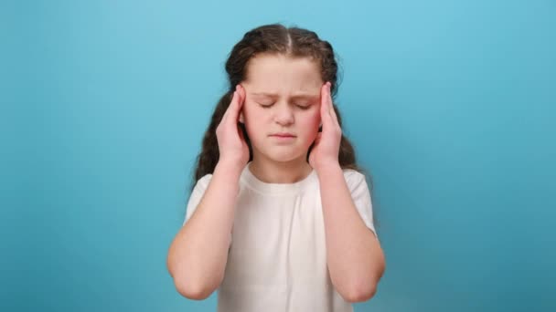 照片中不幸的小女孩紧紧抓住太阳穴 感到极度头痛 儿童偏头痛 健康问题 穿着白色T恤 在工作室的蓝色背景墙上摆出孤立的姿势 — 图库视频影像