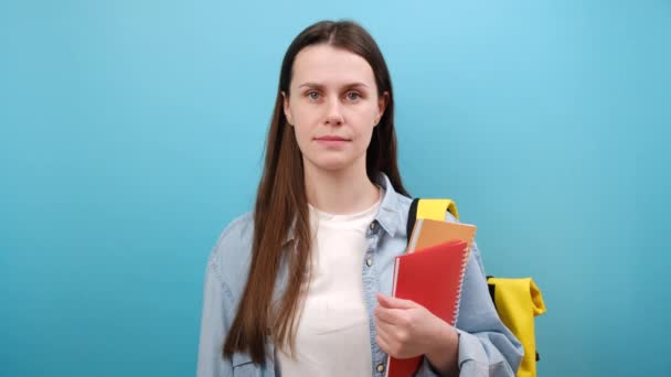 穿着衬衫 背着背包的快乐的女学生的画像 手里拿着两本书和一个大的黄色标签 被隔离在工作室的蓝色背景墙上 社会网络监测和教育的概念 — 图库视频影像