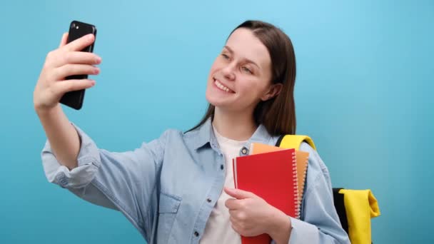 身穿衬衫 背着书包的可爱女学生的肖像 自拍手机后的照片 在蓝色的工作室背景下显得孤立无援 高中教育的概念 — 图库视频影像