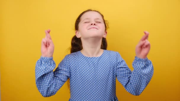 青いドレスを着て8 9歳の少女の肖像画は 特別な瞬間を待って 指を交差させ 願いを作り 目を閉じ スタジオの黄色い背景の壁の上に隔離しました 子供時代のコンセプト — ストック動画