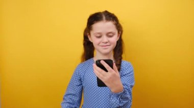 8-9 yaşlarındaki mavi elbiseli küçük kız çocuğunun portresi cep telefonu tutarken sarı arka plan duvarında izole edilmiş bir şekilde el hareketini gösteriyor. Çocukluk yaşam tarzı konsepti