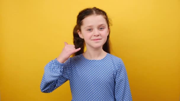 携帯電話のジェスチャーをしている青いドレスで8 9歳の少女の笑顔の肖像画は スタジオで黄色の背景の壁の上に隔離された私を呼び戻すと言います 子供時代のライフスタイルコンセプト — ストック動画