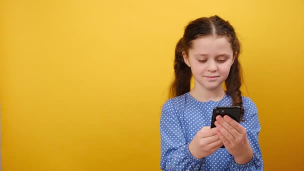 9岁女孩身穿蓝色连衣裙 头脑清醒地梦寐以求的可爱小女孩的画像 把手机放在一边看 在工作室的黄色背景墙上显得孤立无援 童年生活方式概念 — 图库视频影像