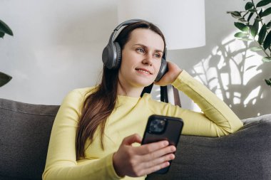 Rahat, esmer, kulaklık takan, akıllı telefon uygulaması ile müzik dinleyen, koltukta dinlenen, elinde cep telefonuyla podcast yapan güzel bir kız.